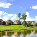 Uma vista encantadora de casas elegantes à beira de um lago tranquilo, cercadas por grama verde exuberante e um céu azul claro com nuvens brancas esparsas, representando um ambiente idílico para concretizar sonhos de vida pacífica e próspera.