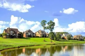 Uma vista encantadora de casas elegantes à beira de um lago tranquilo, cercadas por grama verde exuberante e um céu azul claro com nuvens brancas esparsas, representando um ambiente idílico para concretizar sonhos de vida pacífica e próspera.