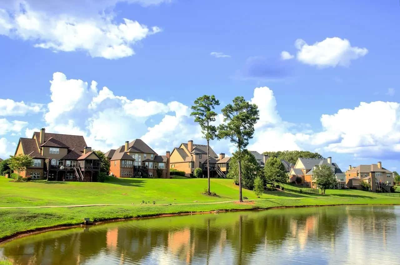 Uma vista encantadora de casas elegantes à beira de um lago tranquilo, cercadas por grama verde exuberante e um céu azul claro com nuvens brancas esparsas, representando um ambiente idílico para concretizar sonhos de vida pacífica e próspera.