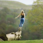 Mulher confiante em pé sobre tronco de árvore em campo aberto, simbolizando liberdade e a busca por sonhos pessoais, ideal para conteúdos relacionados a motivação e resiliência.