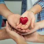 Várias mãos de diferentes pessoas entrelaçadas formando um círculo, simbolizando a união e a força coletiva da solidariedade. O fundo tem um gradiente de cores quentes que representa apoio e inclusão.