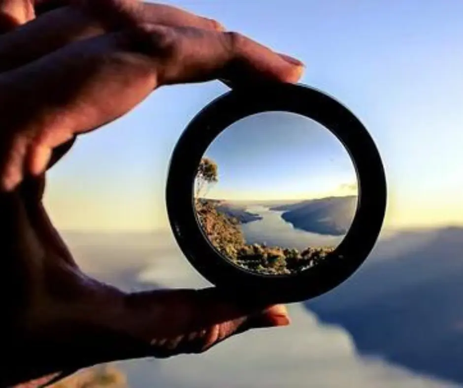 Fotografia inspiradora mostrando uma mão segurando uma lente que reflete um belo cenário natural ao pôr do sol, simbolizando foco e clareza na busca por sucesso e resiliência na vida cotidiana – perfeita para ilustrar artigos sobre superação e atitudes vencedoras como “Ouro na Pista e na Vida: Resiliência Olímpica