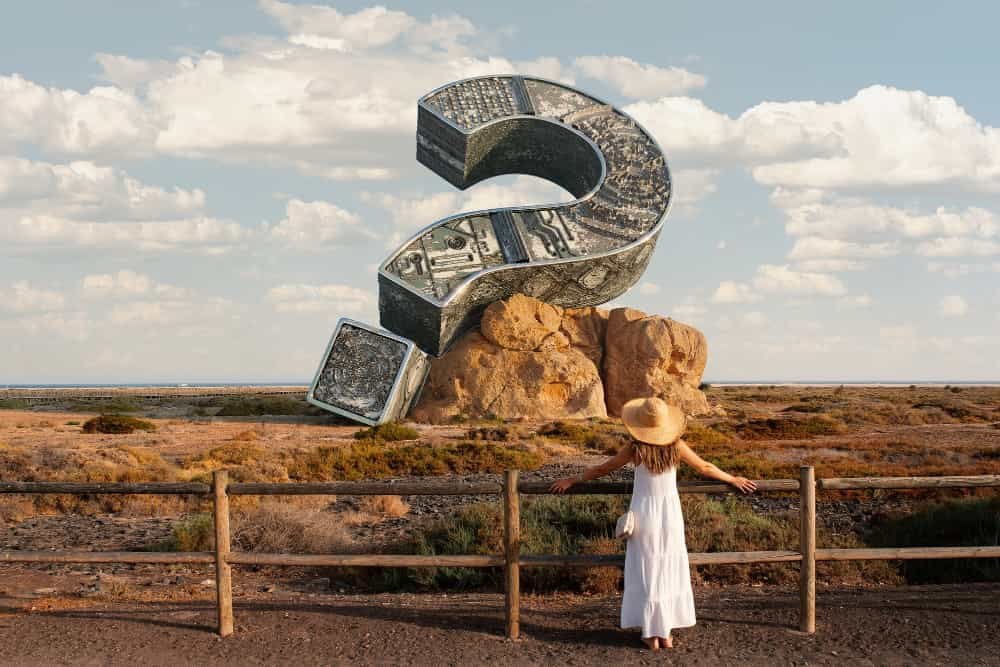 A imagem retrata uma cena surreal onde uma pessoa observa um grande ponto de interrogação feito de notas de dólar, contra o pano de fundo de uma paisagem árida. Elementos específicos incluem: Ponto de Interrogação Gigante: Dominando a cena, parece ser construído a partir de seções de notas de dólar ampliadas, com os designs intrincados e a tonalidade esverdeada claramente visíveis. Pessoa: De pé no primeiro plano, de costas para a câmera, vestindo um vestido branco e um chapéu de abas largas, observando o grande ponto de interrogação. Paisagem: Um vasto terreno árido com grama seca e arbustos esparsos, sob um céu azul claro com nuvens esparsas. Cerca de Madeira: Uma cerca rústica de madeira corre horizontalmente pela imagem, à frente da pessoa. Esta imagem mostra uma justaposição surreal de uma pessoa observando um grande ponto de interrogação feito de notas de dólar em uma paisagem árida, evocando temas de incerteza financeira ou questões sobre riqueza.