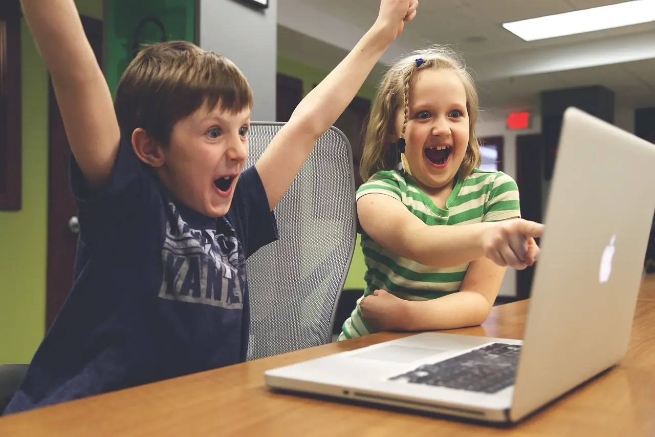 Duas pessoas interagem com entusiasmo em frente a um laptop, uma apontando para a tela e a outra com a mão erguida em um gesto de realização.