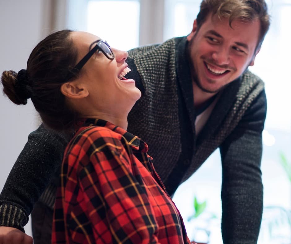 Duas pessoas colaborando em um ambiente de trabalho, com uma delas sentada em frente a um laptop e a outra em pé, oferecendo suporte e orientação. Esta imagem ilustra a importância da cooperação e do trabalho em equipe para aumentar a produtividade e a eficiência no trabalho. A troca de ideias e o apoio mútuo são essenciais para tomar decisões profissionais sábias.
