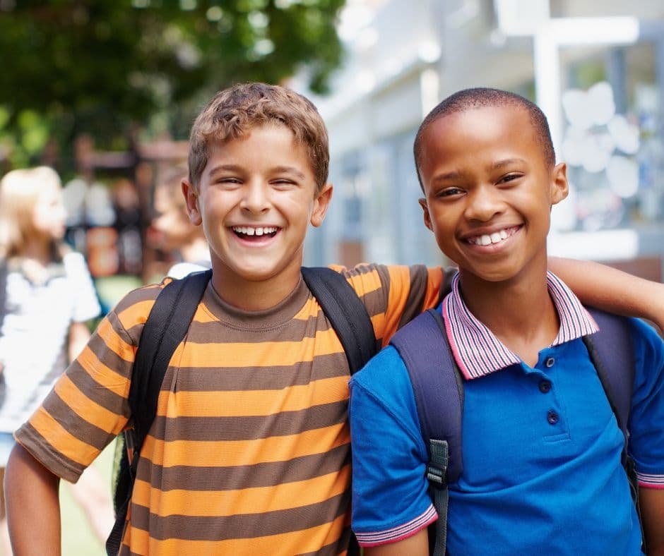 Dois amigos de braços dados, usando mochilas, em um ambiente escolar ao ar livre. A imagem ilustra a importância da amizade e do apoio mútuo no desenvolvimento pessoal e emocional, destacando a relevância de relacionamentos positivos para alcançar o sucesso.