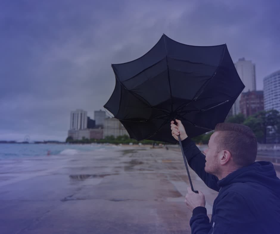 Pessoa segurando um guarda-chuva preto virado ao avesso em uma praia urbana, com prédios altos ao fundo e céu nublado, representando superação de desafios. Imagem do artigo "Supere Seus Obstáculos: O Início", da categoria "Supere Seus Obstáculos". Ideal para conteúdos sobre resiliência, determinação e estratégias para enfrentar adversidades e seguir rumo ao sucesso.